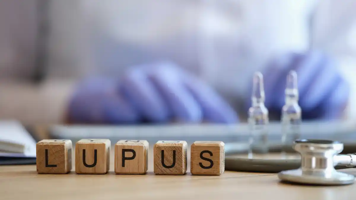 Das Wort Lupus ist auf Holzblöcken zu sehen, daneben liegt ein Stethoskop und 2 Ampullen dahinter, hinten im Hintergrund sieht man unscharf eine Person an einer Tastatur sitzen.