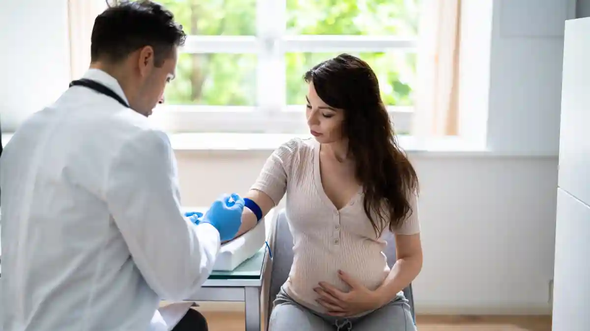 Auf dem Bild sitzt eine junge schwangere Frau auf einem Sessel neben einem Arzt, der ihr gerade Blut abnehmen möchte.