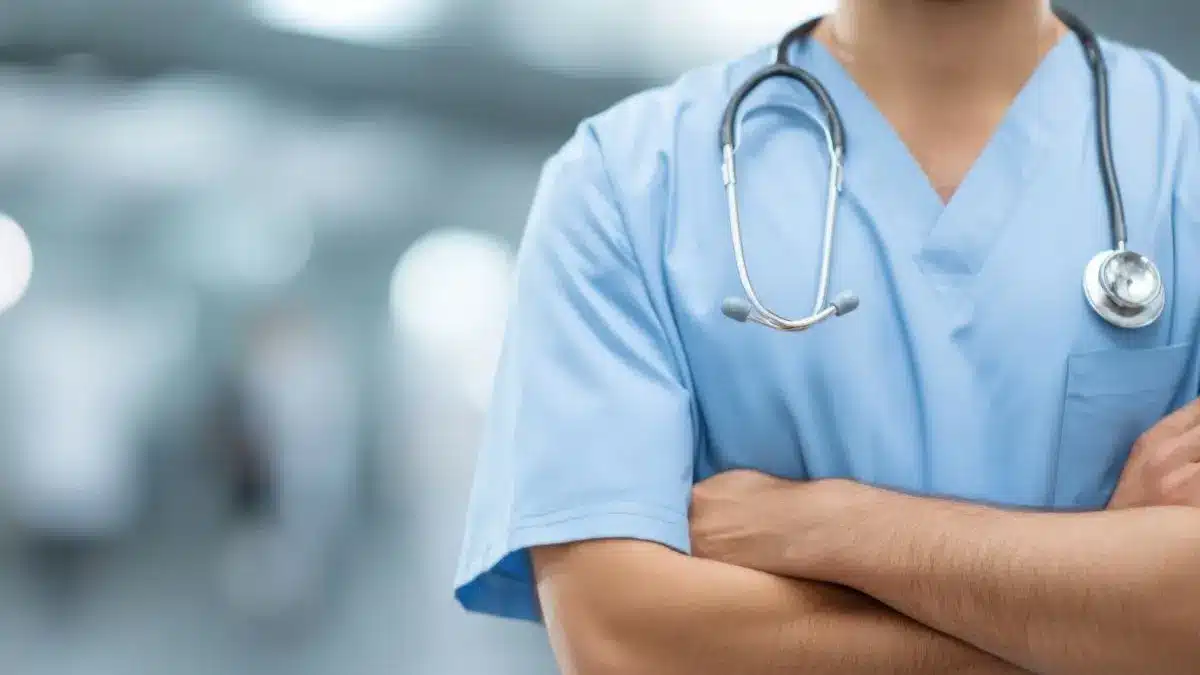 Arzt im blauen Kittel mit verschraenkten Armen und Stethoskop im Klinikflur als Symbol fuer Arbeiten und Weiterbildung in einer Rehabilitationsklinik