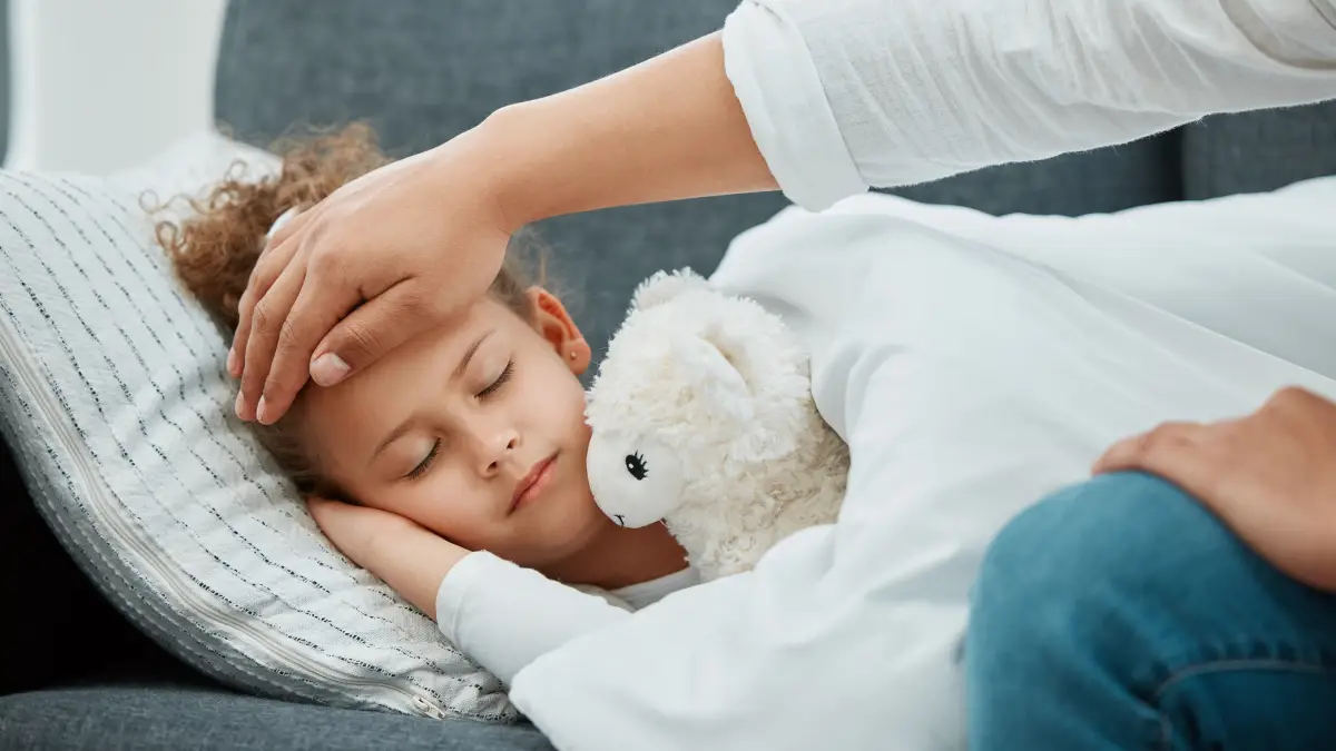 Kleines Mädchen hat Fieber und schläft, erwachsene Person checkt die Temperatur an der Stirn des Kindes mit der Hand
