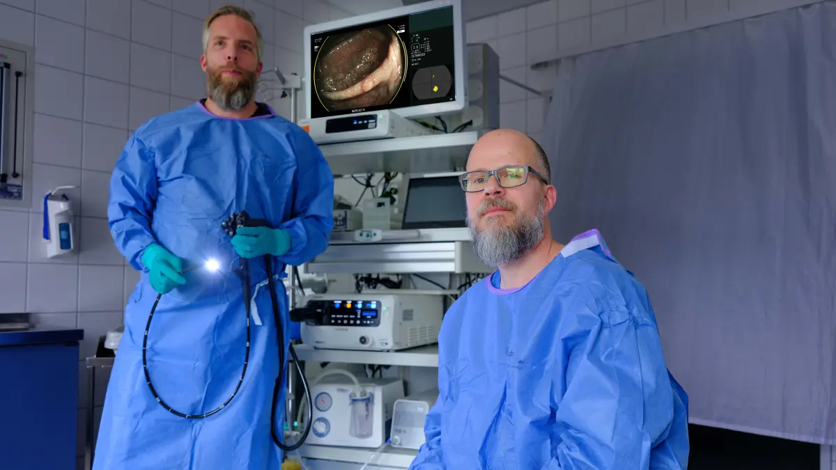Zwei Aerzte in blauer Schutzkleidung in einem Endoskopieraum, einer haelt ein leuchtendes Endoskop, im Hintergrund ist eine Koloskopieaufnahme auf einem Monitor zu sehen