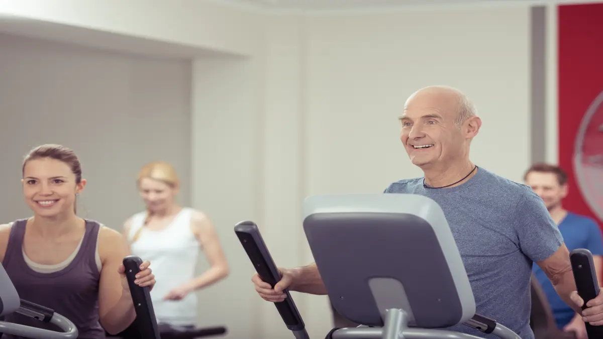 Älterer Mann und junge Frau trainieren lächelnd an Fitnessgeräten im Fitnessstudio – gemeinsames Kraft- und Ausdauertraining