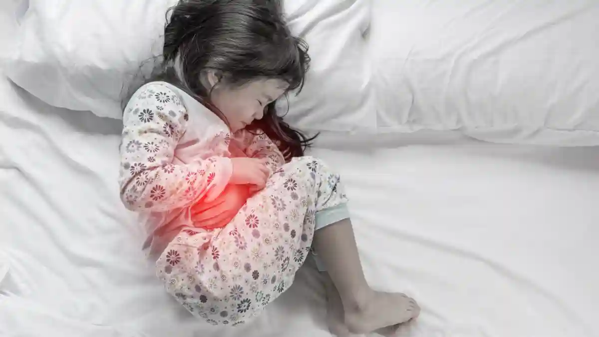 Auf dem Schwarz-Weiß-Bild liegt ein kleines Mädchen auf einem Bett und hält sich den Bauch, der auf dem Bild rot leuchtet.