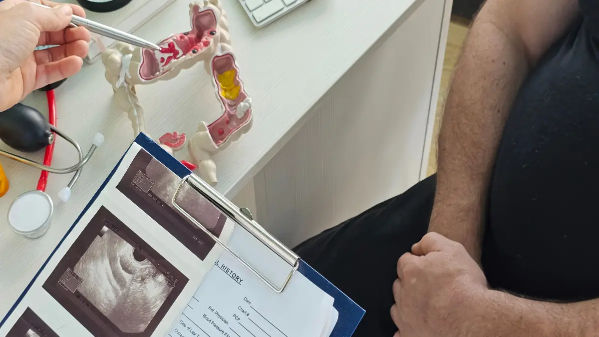 Arzt zeigt mit Stift auf anatomisches Darmmodell mit sichtbarem Tumor während eines Patientengesprächs mit Ultraschallbefunden auf Klemmbrett
