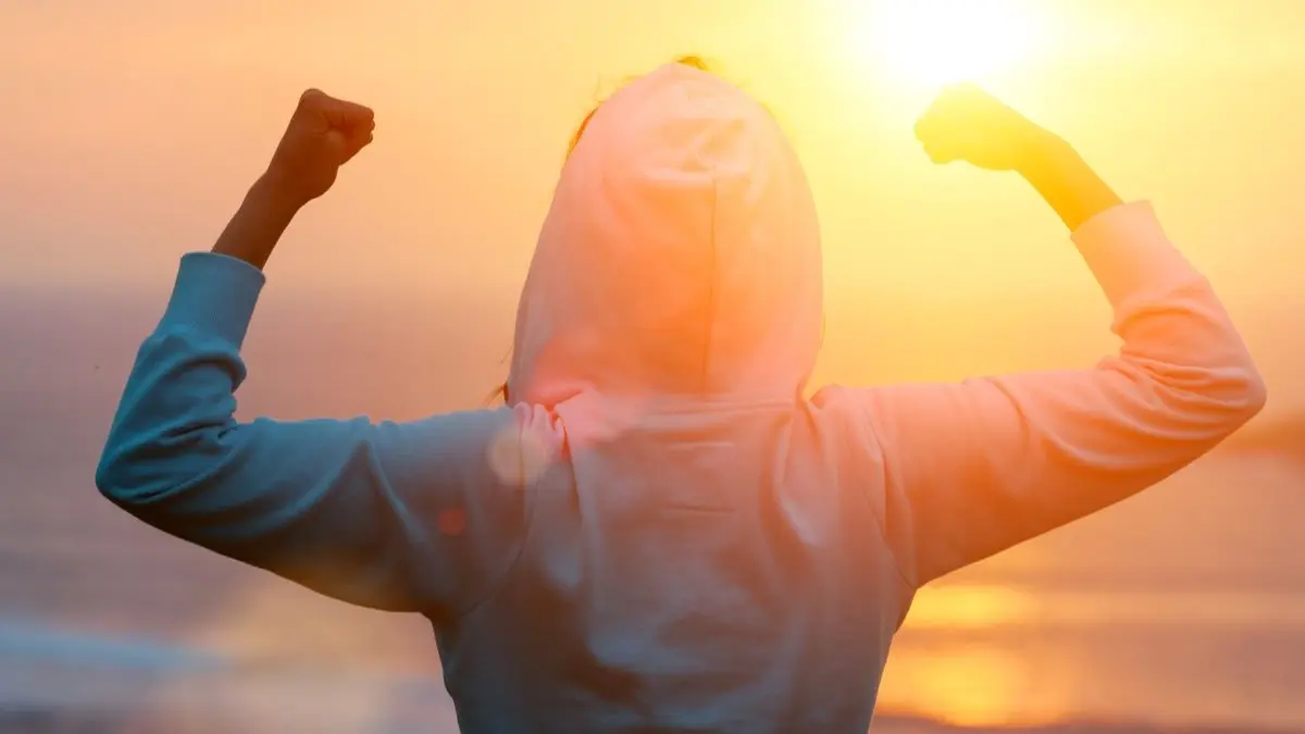 Person mit Kapuzenpulli hebt kraftvoll die Arme vor Sonnenaufgang als Symbol fuer Resilienz bei Stress Leistungsabfall und Hautentzuendungen