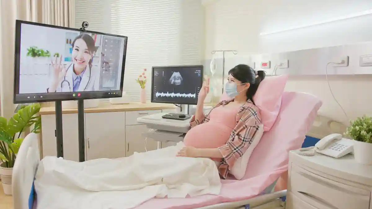 Eine schwangere Frau liegt auf einem Krankenhausbett und bekommt einen Videocall, der über einen Bildschirm übertragen wird, im Bilschirm ist eine Ärztin zu sehen.