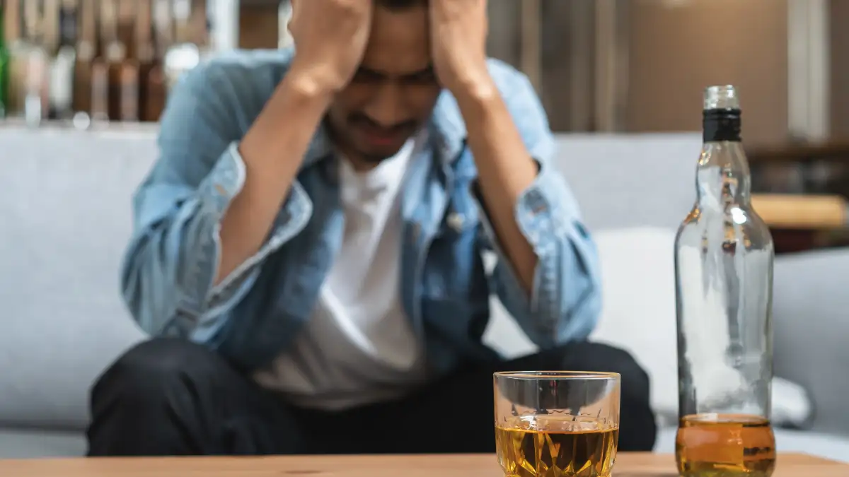 Junger Mann sitzt mit Händen vor der Stirn auf dem Sofa, vor ihm auf dem Couchtisch steht eine Flasche und ein Glas mit Alkohol.