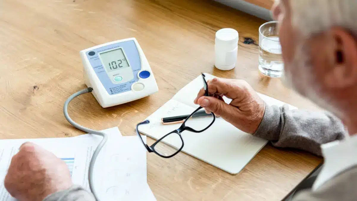 Älterer Mann am Tisch vor einem Blutdruckmessgerät und medizinischen Unterlagen – Blutdruckkontrolle und Medikamentenmanagement im Alter