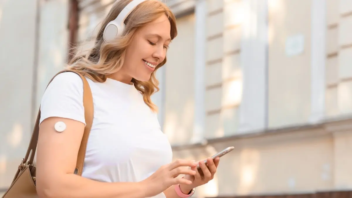 Junge Frau mit Glukosesensor am Oberarm geht laechelnd durch die Stadt und blickt auf ihr Smartphone