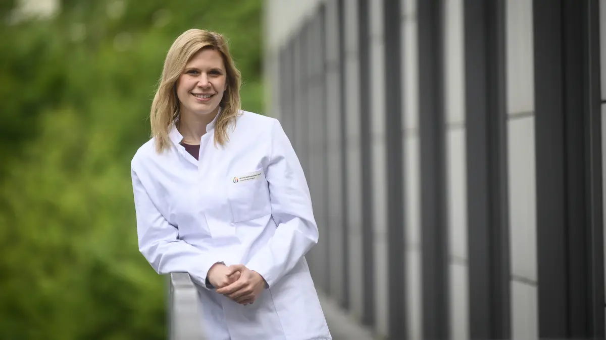 Wissenschaftlerin im weissen Kittel lehnt an einem Gelaender vor einem Klinikgebaeude, Symbol fuer Forschung zu schwarzem Hautkrebs im Emmy-Noether-Programm