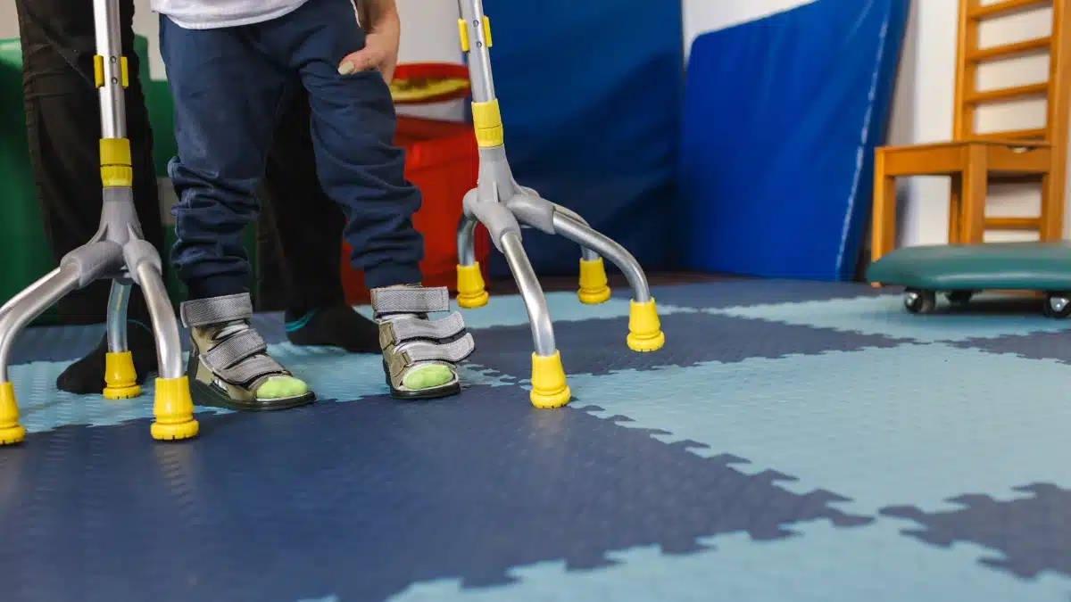 Kleiner Junge mit Bewegungsstörungen übt mir Unterstützung einer Physiotherapeutin das Gehen mit Vierfußgehhilfe