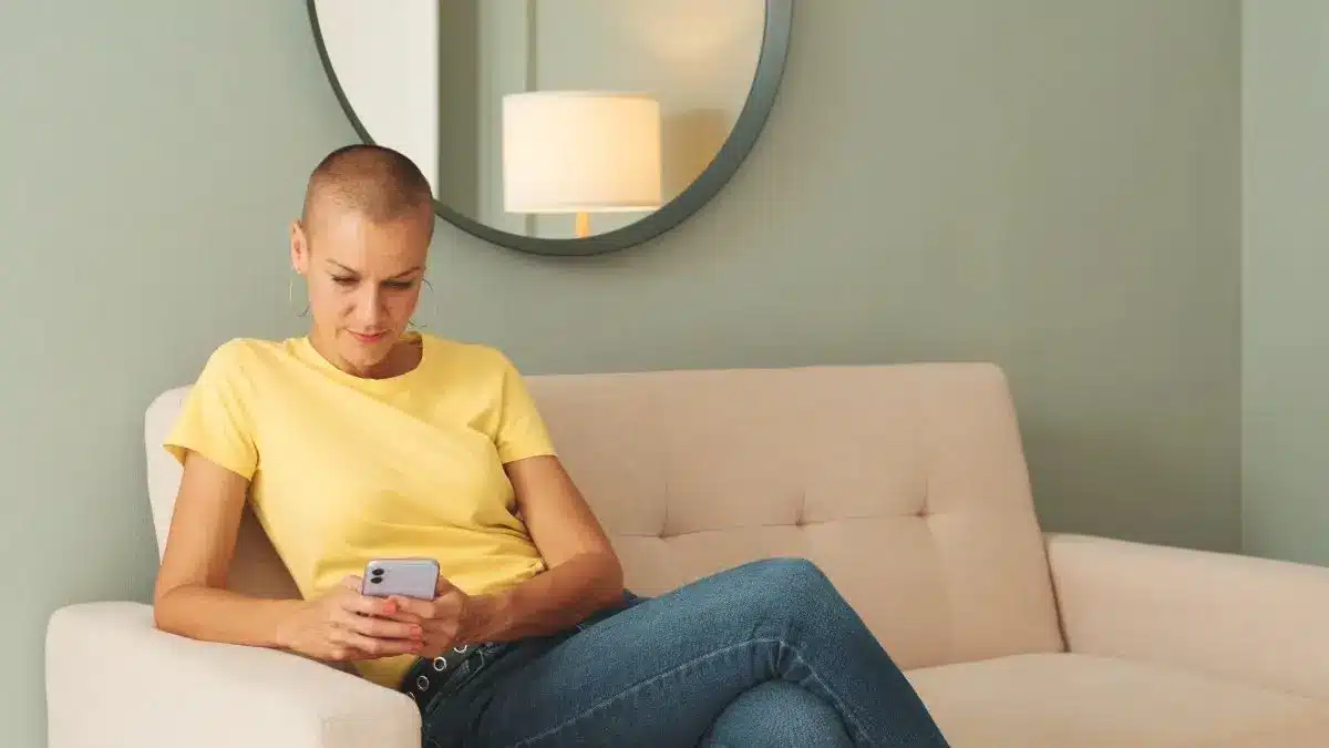 Patientin nach Brustkrebstherapie sitzt auf dem Sofa und nutzt die GenE3 App auf dem Smartphone zur Unterstuetzung bei familiaerem Brust und Eierstockkrebs