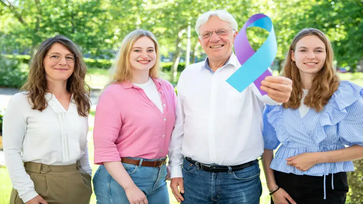 Gruppe von vier Personen im Freien, eine Person haelt eine zweifarbige Krebsbewusstseins-Schleife als Symbol fuer das ROKAVI-Projekt zur KI-gestuetzten Krebsvorhersage mit Krankenkassendaten