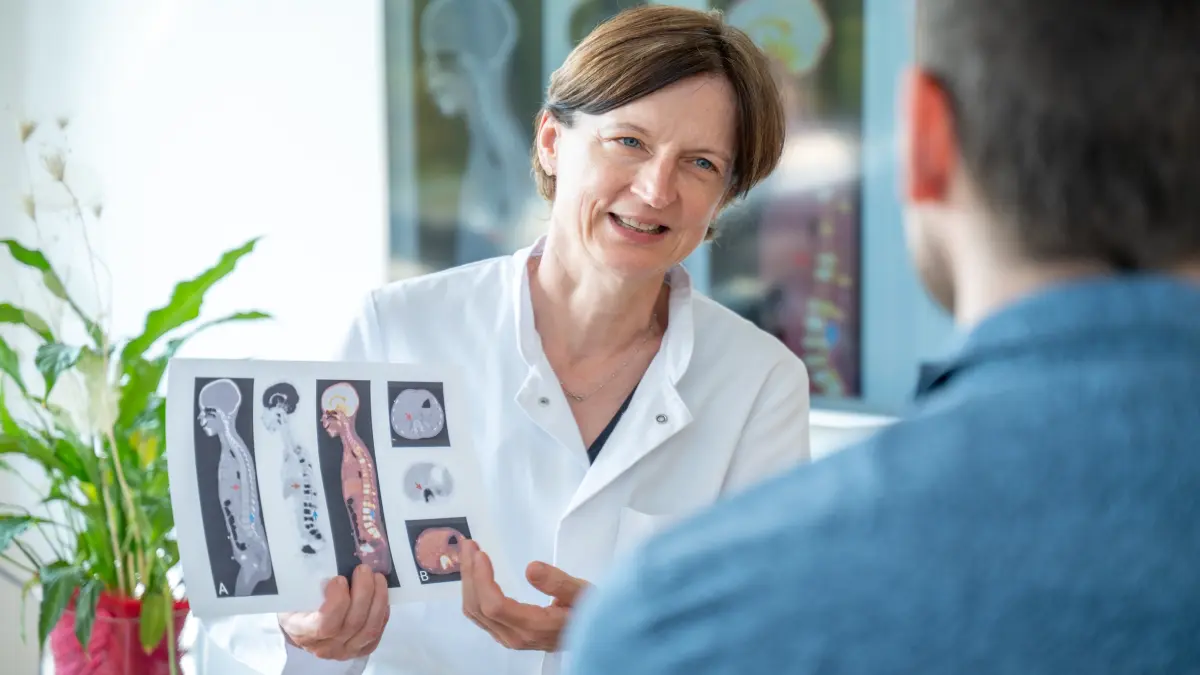 Aerztin im weissen Kittel erklaert einem Patienten PET-Scan-Bilder als schonende Alternative zur schmerzhaften Knochenmarkprobe bei Muskelkrebs Rhabdomyosarkom
