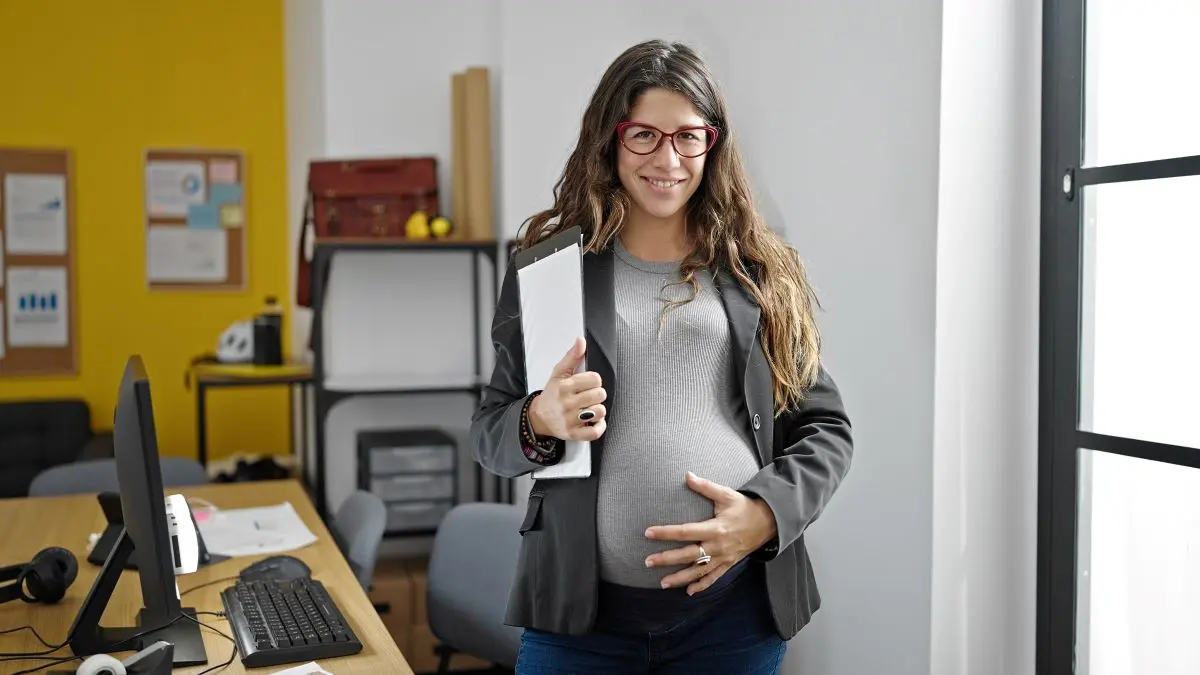 Schwangere selbstaendige Unternehmerin steht mit Aktenmappe im Buero und haelt die Hand auf den Babybauch als Symbol fuer Mutterschaftsausgleich fuer Selbstaendige