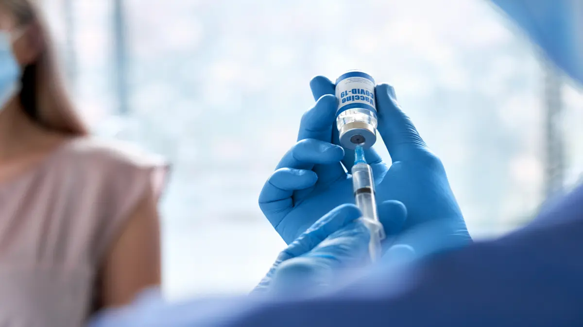 Arzt in blauen Schutzhandschuhen zieht COVID-19-Impfstoff aus einem Fläschchen in eine Spritze auf, im Hintergrund eine Patientin