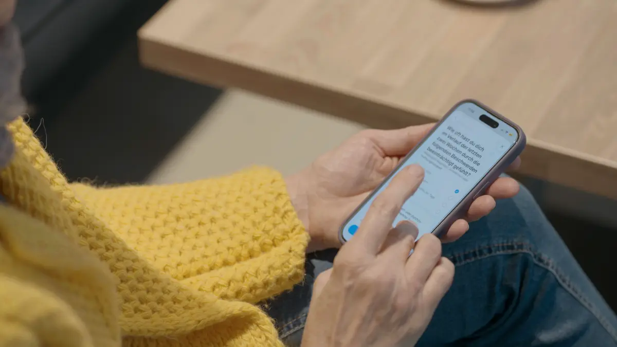 Person mit gelbem Strickpulli bedient eine App auf dem Smartphone, die Fragen zu Symptomen und Befinden stellt