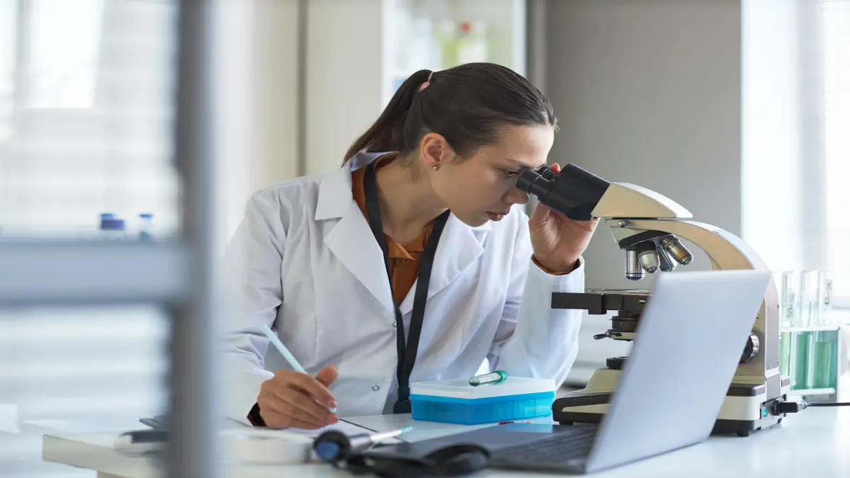 Wissenschaftlerin im Laborkittel schaut durch ein Mikroskop und macht Notizen