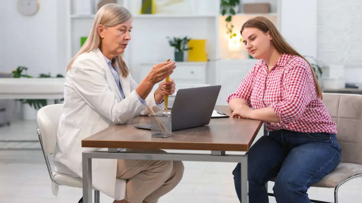 Eine ältere Ärztin sitzt an einem Tisch mit einem aufgeklappten Computer, eine Patientin sitzt ihr gegenüber.