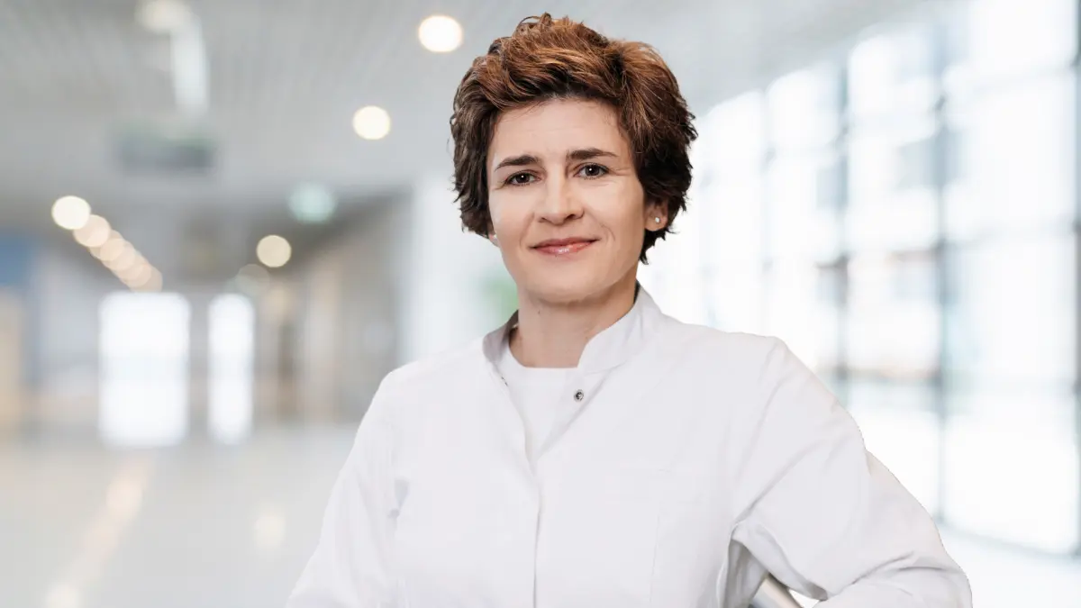 Aerztin in weissem Kittel steht in einem hellen Klinikflur und laechelt in die Kamera, Symbolbild fuer die Leitung eines Forschungsinstituts zur akuten lymphoblastischen Leukaemie