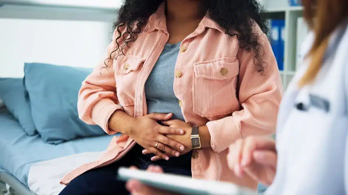 Junge Frau sitzt in einer Praxis auf dem Bett und haelt sich mit beiden Haenden den Unterbauch waehrend eine Aerztin gegenuebersitzt
