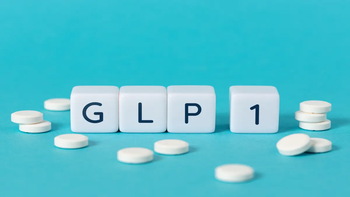 Weiße Würfel mit der Aufschrift „GLP-1“ und verstreute weiße Tabletten auf türkisfarbenem Hintergrund.