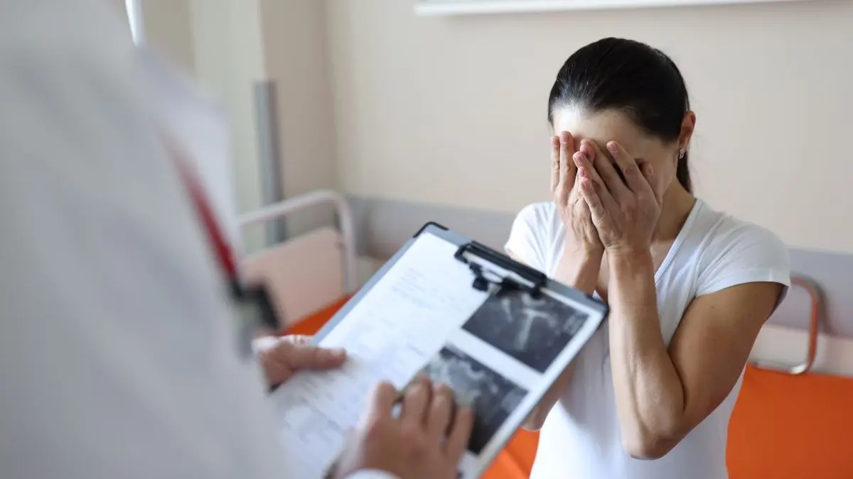 Frau haelt sich im Krankenhauszimmer das Gesicht in den Haenden waehrend ein Arzt vor ihr mit Klemmbrett und Ultraschallbildern steht