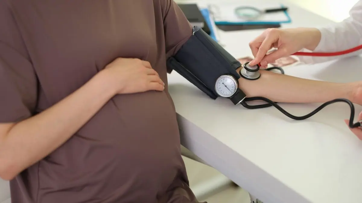 Schwangere Frau sitzt am Tisch, haelt eine Hand auf den Bauch, waehrend an ihrem Arm der Blutdruck gemessen wird