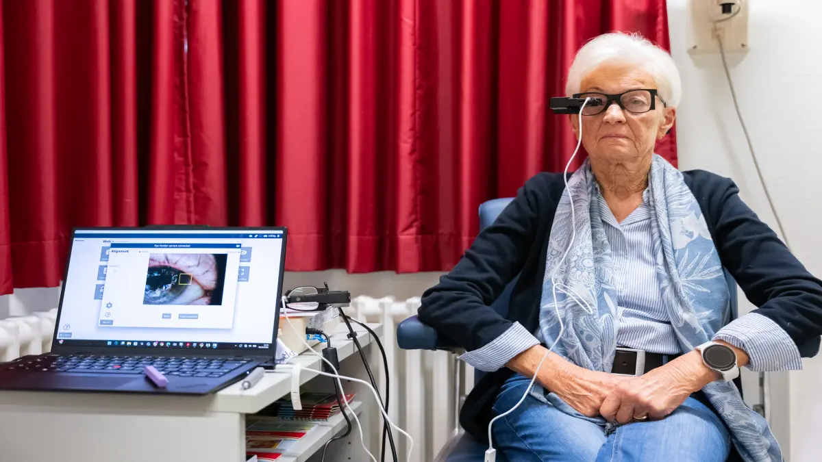 Ältere Patientin mit weißen Haaren und Brille trägt eine Spezialbrille mit Mikrokamera, daneben ein Laptop mit Einstellungsbildschirm