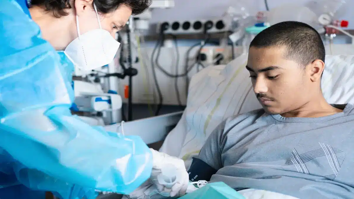 Aerztin in Schutzkleidung und FFP2-Maske verabreicht einem jungen Patienten im Krankenhausbett eine Infusion mit genveraenderten Stammzellen