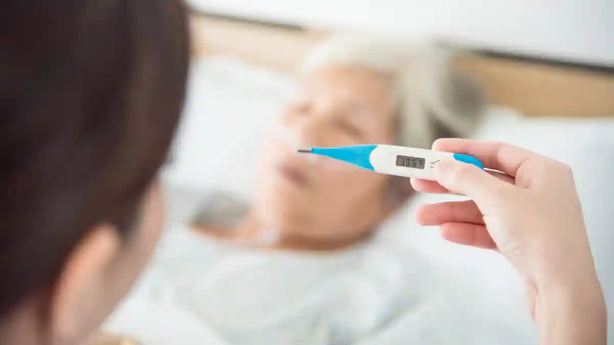 Auf dem Foto hält eine Krankenschwester ein Thermometer mit hohem Temperaturergebnis dahinter sieht man verschwommen eine ältere Frau in einem Bett.