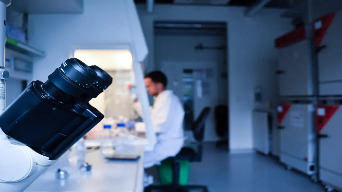 Schwarzes Labormikroskop im Vordergrund, im Hintergrund arbeitet ein Wissenschaftler im weißen Kittel an einem Labortisch