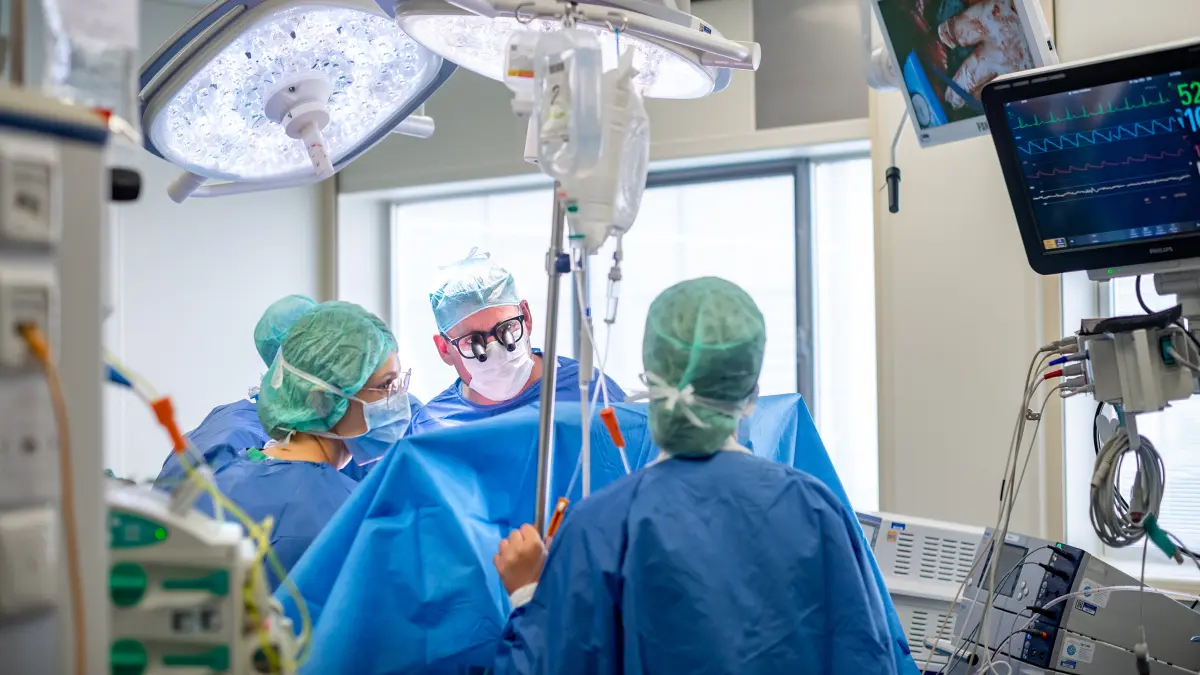 Operationsteam in blauen OP-Kitteln und gruener Haube fuehrt unter hellem OP-Licht eine Lebertransplantation durch, im Vordergrund sind Monitore und Infusionen zu sehen
