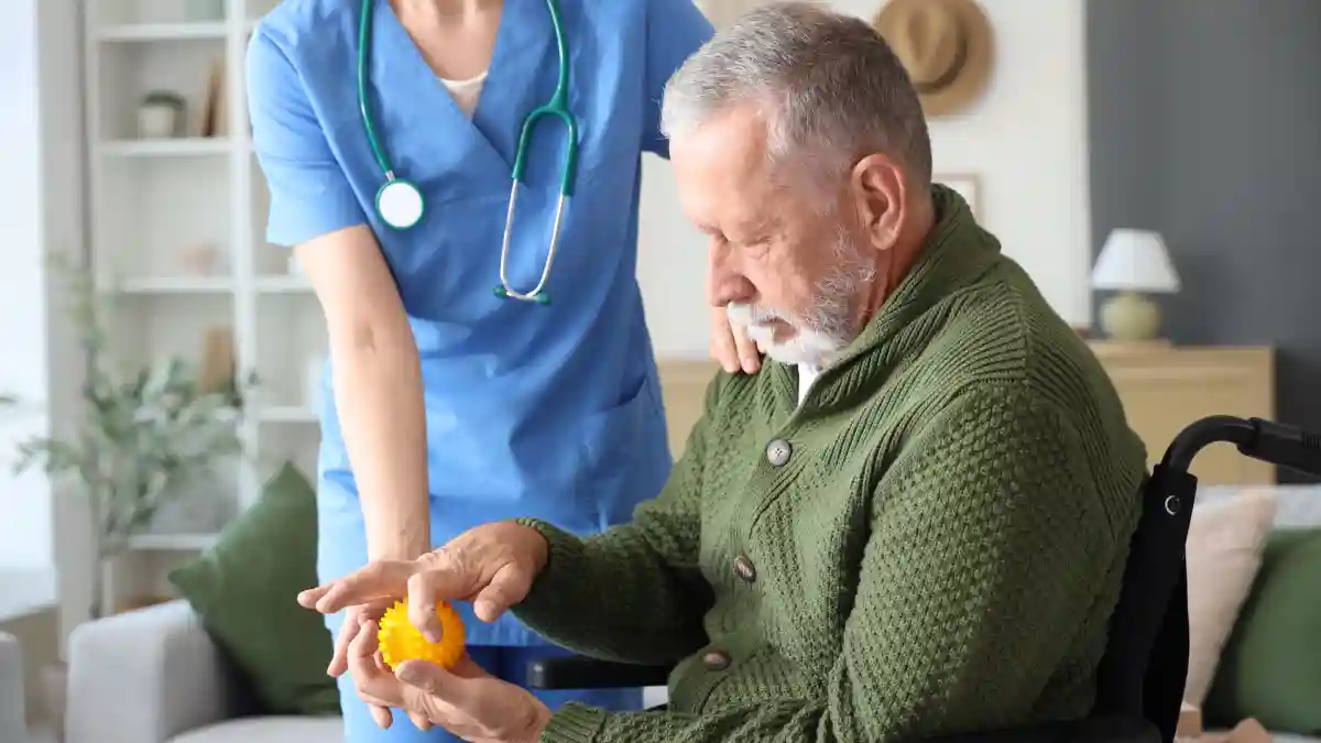 Auf dem Bild sitzt ein älterer Mann in einem Rollstuhl, in den Händen hält er einen gelben Massageball, neben ihm ist eine Krankenschwester.